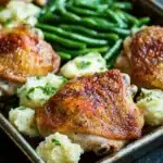Chicken thigh dinner and sides - the image shows a rectangular baking dish filled with roasted chicken and green beans. the chicken is golden brown and appears to be seasoned with herbs and spices. the green beans are bright green and look fresh and vibrant. the dish is garnished with chopped chives, adding a pop of color to the dish. the background is a dark grey countertop.