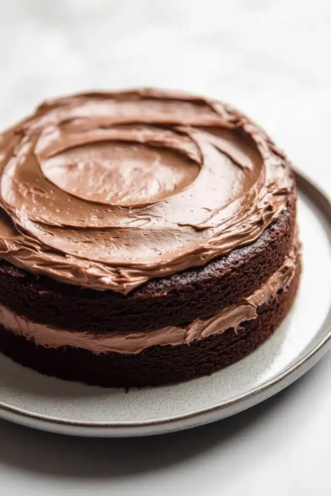 Chocolate cake normal design - the image is of a round chocolate cake on a white plate. the cake appears to be freshly baked and has a smooth texture. the top of the cake is covered in a generous layer of chocolate frosting, which is spread evenly across the cake. the frosting is a rich, dark brown color and looks smooth and glossy. the plate is round and has small speckles around the edges. the background is blurred, making the cake the focal point of the image.