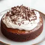 Chocolate cake tres leches - the image is of a round chocolate cake on a white plate. the cake appears to be freshly baked and has a rich, dark chocolate color. on top of the cake, there is a generous dollop of whipped cream and sprinkled with chocolate shavings. the plate is sitting on a marble countertop with a cup of coffee in the background.