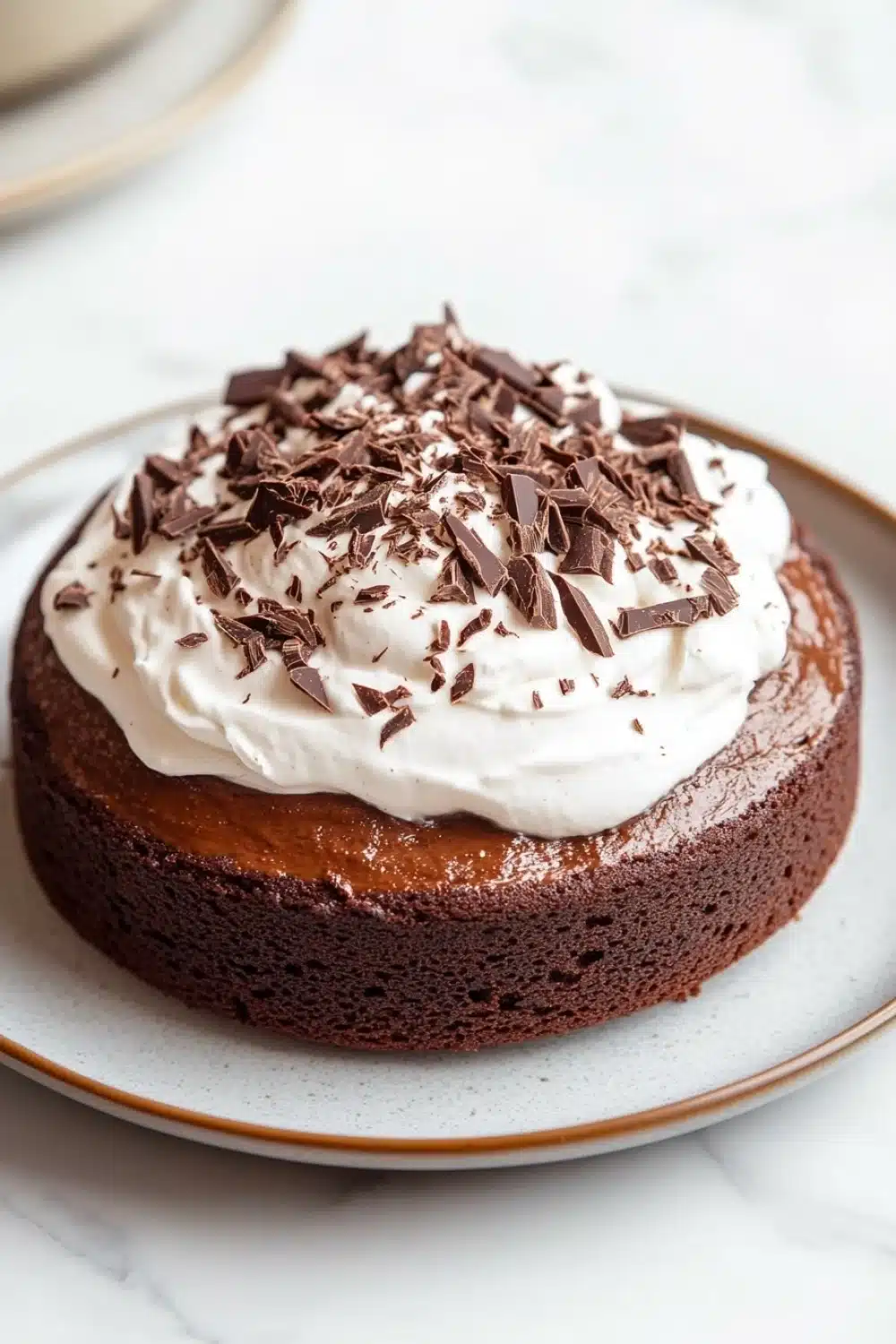 Chocolate cake tres leches - the image is of a round chocolate cake on a white plate. the cake appears to be freshly baked and has a rich, dark chocolate color. on top of the cake, there is a generous dollop of whipped cream and sprinkled with chocolate shavings. the plate is sitting on a marble countertop with a cup of coffee in the background.