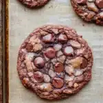Chocolate chip cookie no chocolate chips - the image shows three freshly baked chocolate chip cookies on a baking tray. the cookies are round and golden brown in color, with chocolate chips scattered throughout. they are arranged in a triangular formation on the tray, with the largest cookie in the center and two smaller cookies on either side. the tray is lined with parchment paper, and the cookies appear to be freshly baked. the background is a wooden table.