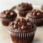 Chocolate muffin vegan - the image shows a group of freshly baked chocolate muffins on a white baking sheet. the muffins are round and have a golden brown color. they are topped with chocolate chips and drizzled with chocolate sauce. the tray is lined with parchment paper and there are a few more muffins scattered around the tray. the background is blurred, but it appears to be a kitchen countertop.