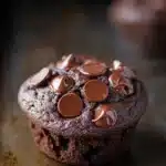 Chocolate muffin without eggs - the image is a close-up of a chocolate muffin on a baking tray. the muffin is round and has a golden brown color. it is topped with chocolate chips that are arranged in a circular pattern. the chocolate chips are evenly spaced and appear to be freshly baked. the tray is lined with parchment paper and there are a few more muffins visible in the background. the overall mood of the image is warm and inviting.
