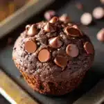Chocolate quinoa muffin - the image shows a chocolate muffin on a baking tray. the muffin is round and has a golden brown color. it is covered in chocolate shavings, which are scattered across the top of the muffin. there are also small pieces of chocolate scattered around the tray, some of which are also covered in the same chocolate. the tray is sitting on a wooden surface, and there are a few more chocolate chips visible in the background.