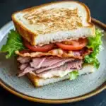 Club sandwich beef - the image is a close-up of a sandwich on a plate. the sandwich is made with two slices of toasted bread, filled with sliced ham, bacon, and lettuce. the ham is pink and appears to be seasoned with herbs and spices. the bacon is crispy and golden brown, and the lettuce is a vibrant green color. the plate is a light blue color with a speckled pattern. the background is dark, making the sandwich stand out.