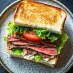 Club sandwich beef - the image shows a sandwich on a plate. the sandwich is made with two slices of toasted bread, filled with sliced beef, lettuce, and a tomato. the beef appears to be cooked medium-rare, with a pinkish-red color and a slightly charred texture. the lettuce is a bright green color, and the tomato is a deep red. the plate is a light blue color with a speckled pattern. the background is dark, making the sandwich stand out.