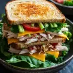 Club sandwich ensalada de pollo - the image shows a sandwich in a black bowl. the sandwich is made with two slices of toasted bread, filled with shredded chicken, lettuce, tomato, and cheese. the chicken appears to be seasoned with herbs and spices, and the cheese is melted and oozing out of the sides. the lettuce is a mix of green and yellow, and there are a few slices of red onion scattered throughout the sandwich. in the background, there is a small bowl of salad. the bowl is sitting on a dark countertop.