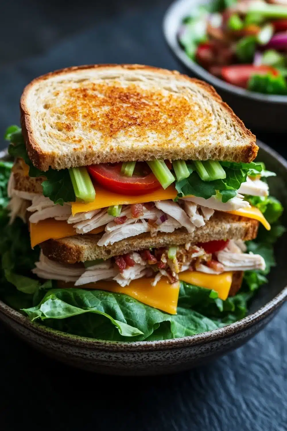 Club sandwich ensalada de pollo - the image shows a sandwich in a black bowl. the sandwich is made with two slices of toasted bread, filled with shredded chicken, lettuce, tomato, and cheese. the chicken appears to be seasoned with herbs and spices, and the cheese is melted and oozing out of the sides. the lettuce is a mix of green and yellow, and there are a few slices of red onion scattered throughout the sandwich. in the background, there is a small bowl of salad. the bowl is sitting on a dark countertop.