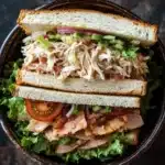Club sandwich ensalada de pollo - the image shows a sandwich in a black bowl. the sandwich is made with two slices of white bread, filled with shredded chicken, lettuce, tomato, and red onion. the chicken appears to be seasoned with herbs and spices, and the lettuce is a mix of green and red colors. the tomato is cut in half and is placed on top of the chicken. the bowl is sitting on a dark countertop.