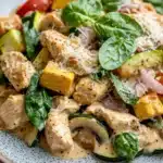 Creamy chicken and vegetable skillet - the image is a close-up of a plate of food. the plate is white with a speckled pattern and is placed on a wooden table. the food appears to be a salad with grilled chicken, zucchini, red bell peppers, and spinach. the chicken is cooked to a golden brown color and is garnished with grated parmesan cheese. the vegetables are cut into small cubes and are mixed together with a creamy sauce. the dish looks fresh and appetizing.