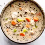 Creamy chicken wild rice soup crockpot - the image is a close-up of a bowl of soup. the soup appears to be creamy and has chunks of chicken and vegetables mixed in. the bowl is made of ceramic and is sitting on a white marble countertop. there are also some sprigs of parsley scattered around the bowl. the overall color scheme of the image is warm and inviting.