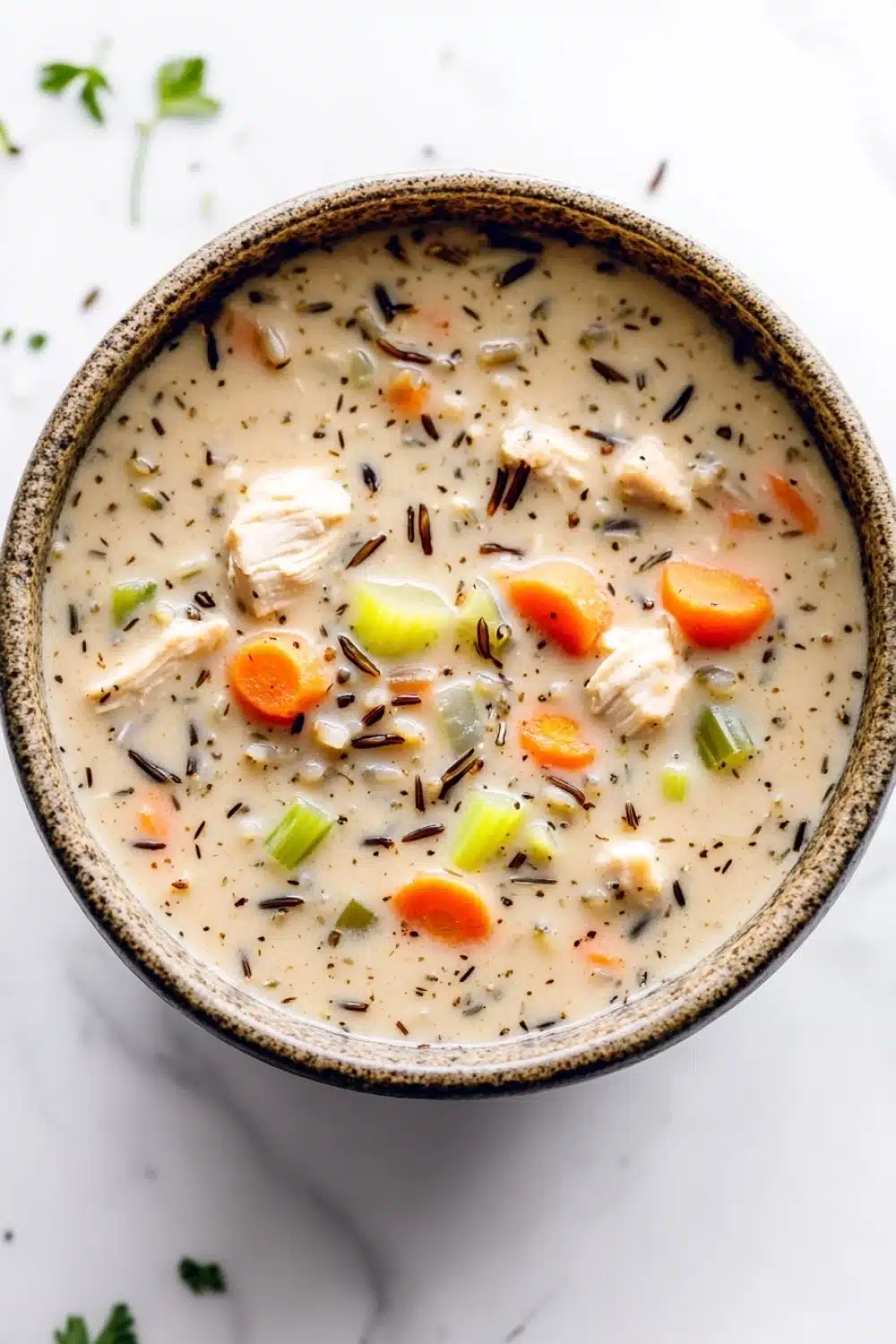 Creamy chicken wild rice soup crockpot - the image is a close-up of a bowl of soup. the soup appears to be creamy and has chunks of chicken and vegetables mixed in. the bowl is made of ceramic and is sitting on a white marble countertop. there are also some sprigs of parsley scattered around the bowl. the overall color scheme of the image is warm and inviting.