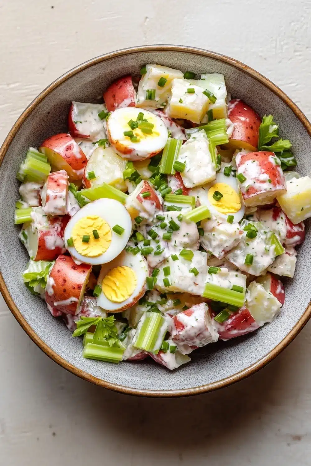 Creamy hot potato salad - the image is a close-up of a bowl of potato salad. the bowl is made of a light-colored ceramic material and is placed on a white countertop. the salad is made up of diced potatoes, diced green onions, and hard-boiled eggs. the potatoes are arranged in a circular pattern, with some overlapping each other. the eggs are cut into small cubes and are scattered throughout the salad. there are also some chopped green herbs sprinkled on top. the overall appearance of the salad is fresh and appetizing.