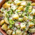 Creamy hot potato salad - the image is a close-up of a bowl of potato salad. the bowl is made of terracotta and is sitting on a white wooden table. the salad is made up of diced potatoes, red onions, green onions, and chopped herbs. the potatoes are cut into small cubes and are arranged in a pile. the herbs are scattered throughout the salad, adding a pop of green color to the dish. the overall appearance of the salad is fresh and appetizing.