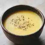 Creamy yellow tomato soup - the image is a close-up of a small bowl of soup. the bowl is made of dark brown ceramic and has a smooth texture. the soup appears to be a creamy yellow color and is topped with small black speckles. the background is a wooden table with a light-colored surface. the overall mood of the image is warm and inviting.