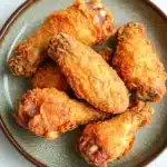 Crispy chicken wings fried - the image shows a plate of fried chicken wings. the wings are golden brown and appear to be crispy on the outside. they are arranged in a circular pattern on the plate, with some overlapping each other. the plate is made of a light-colored ceramic with a speckled texture. the background is a white marble countertop.