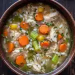 Crock pot chicken rice soup - the image is a close-up of a bowl of soup. the soup appears to be a creamy white color with chunks of chicken, carrots, celery, and other vegetables mixed in. the bowl is made of ceramic and is sitting on a dark wooden surface. there are a few sprigs of parsley scattered around the bowl. the overall appearance of the soup is fresh and appetizing.