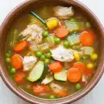 Crock pot chicken vegetable soup - the image is a close-up of a bowl of soup. the soup appears to be a vegetable soup with chunks of chicken, carrots, zucchini, peas, and other vegetables mixed in. the bowl is made of ceramic and is sitting on a white wooden surface. there is a spoon in the soup, suggesting that it is ready to be eaten. the broth is a light brown color and looks thick and creamy.