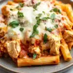 Crockpot chicken ziti slow cooker - the image shows a plate of penne pasta with chicken and tomato sauce. the pasta is cooked al dente and is topped with melted cheese and fresh basil leaves. the dish is served on a gray plate with a white marble countertop. the chicken is cooked to a golden brown color and appears to be seasoned with herbs and spices. the tomato sauce is a deep red color and is drizzled over the top of the pasta. the overall appearance of the dish is savory and appetizing.