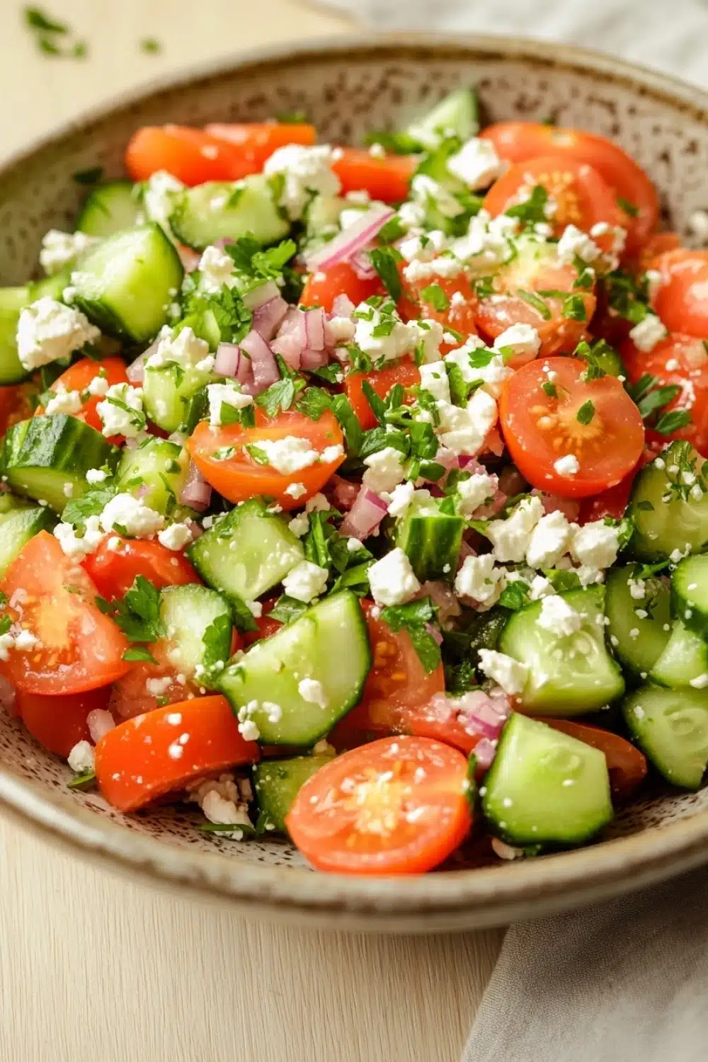Cucumber tomato feta salad - the image is a close-up of a colorful salad in a bowl. the salad is made up of sliced tomatoes, cucumbers, red onions, and feta cheese. the tomatoes are bright red and appear to be fresh, while the cucumbers are green and the onions are red. the feta is crumbled and scattered throughout the salad. the bowl is sitting on a white tablecloth and there are a few sprigs of parsley scattered around the bowl.