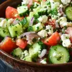 Cucumber tomato feta salad - the image is a close-up of a bowl of salad. the bowl is made of wood and is placed on a dark wooden table. the salad is made up of various ingredients, including sliced cucumbers, cherry tomatoes, red onions, and feta cheese. the cucumbers are cut into thin slices and are arranged in a colorful and appetizing manner. the tomatoes are bright red and appear to be ripe and juicy. the feta is crumbled and scattered throughout the salad, adding a creamy texture. there are also some fresh herbs and cilantro scattered throughout. the overall appearance of the salad is fresh and vibrant.