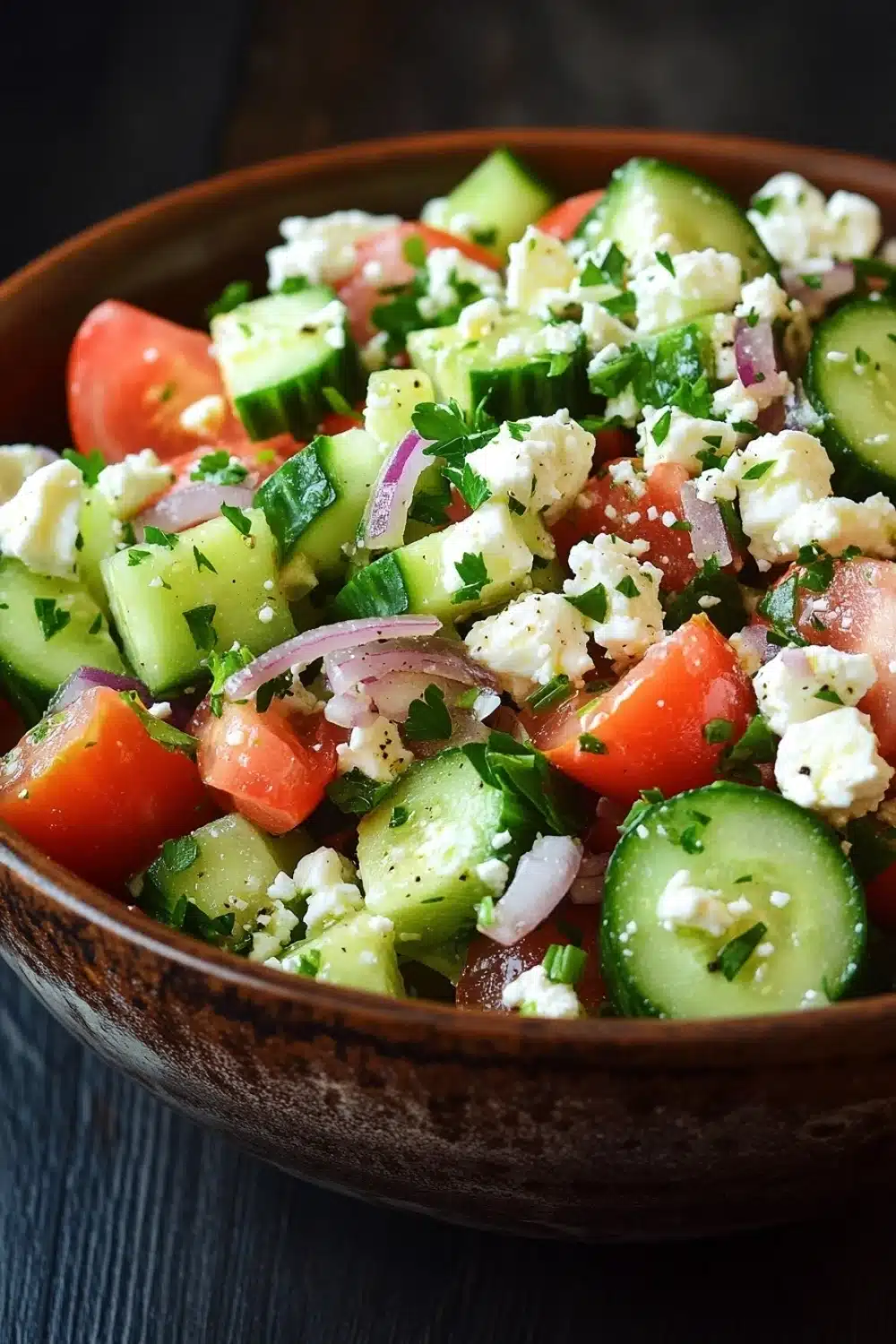 Cucumber tomato feta salad - the image is a close-up of a bowl of salad. the bowl is made of wood and is placed on a dark wooden table. the salad is made up of various ingredients, including sliced cucumbers, cherry tomatoes, red onions, and feta cheese. the cucumbers are cut into thin slices and are arranged in a colorful and appetizing manner. the tomatoes are bright red and appear to be ripe and juicy. the feta is crumbled and scattered throughout the salad, adding a creamy texture. there are also some fresh herbs and cilantro scattered throughout. the overall appearance of the salad is fresh and vibrant.