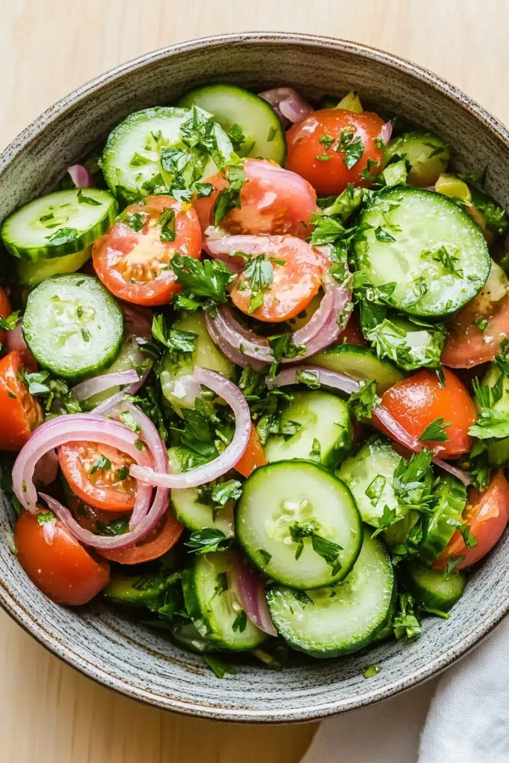 Cucumber tomato onion salad lemon - the image is a close-up of a bowl of salad. the salad is made up of sliced cucumbers, red onions, and cherry tomatoes. the cucumbers are cut into thin slices and are arranged in a circular pattern. the tomatoes are bright red and appear to be ripe and juicy. the onions are sliced into thin strips and are scattered throughout the salad, adding a pop of color to the dish. there are also some fresh herbs, possibly parsley, scattered throughout. the bowl is sitting on a wooden table with a white cloth napkin on the side.