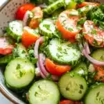 Cucumber tomato onion salad lemon - the image is a close-up of a bowl of salad. the bowl is made of ceramic and has a speckled pattern. the salad is made up of sliced cucumbers, red onions, cherry tomatoes, and chopped herbs. the cucumbers are cut into thin slices and are arranged in a circular pattern around the bowl. the tomatoes are bright red and appear to be ripe and juicy. the herbs are finely chopped and add a pop of green color to the dish. the background is blurred, but it appears to be a kitchen countertop with a sprig of parsley.