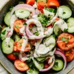 Cucumber tomato onion salad with ranch - the image is a close-up of a bowl of salad. the salad is made up of sliced cucumbers, red onions, cherry tomatoes, and cilantro. the cucumbers are cut into thin slices and are arranged in a circular pattern. the red onions are sliced into thin strips and are scattered throughout the salad. there is also a dollop of white dressing on top of the cucumbers. the bowl is sitting on a white tablecloth with a few sprigs of parsley scattered around it.