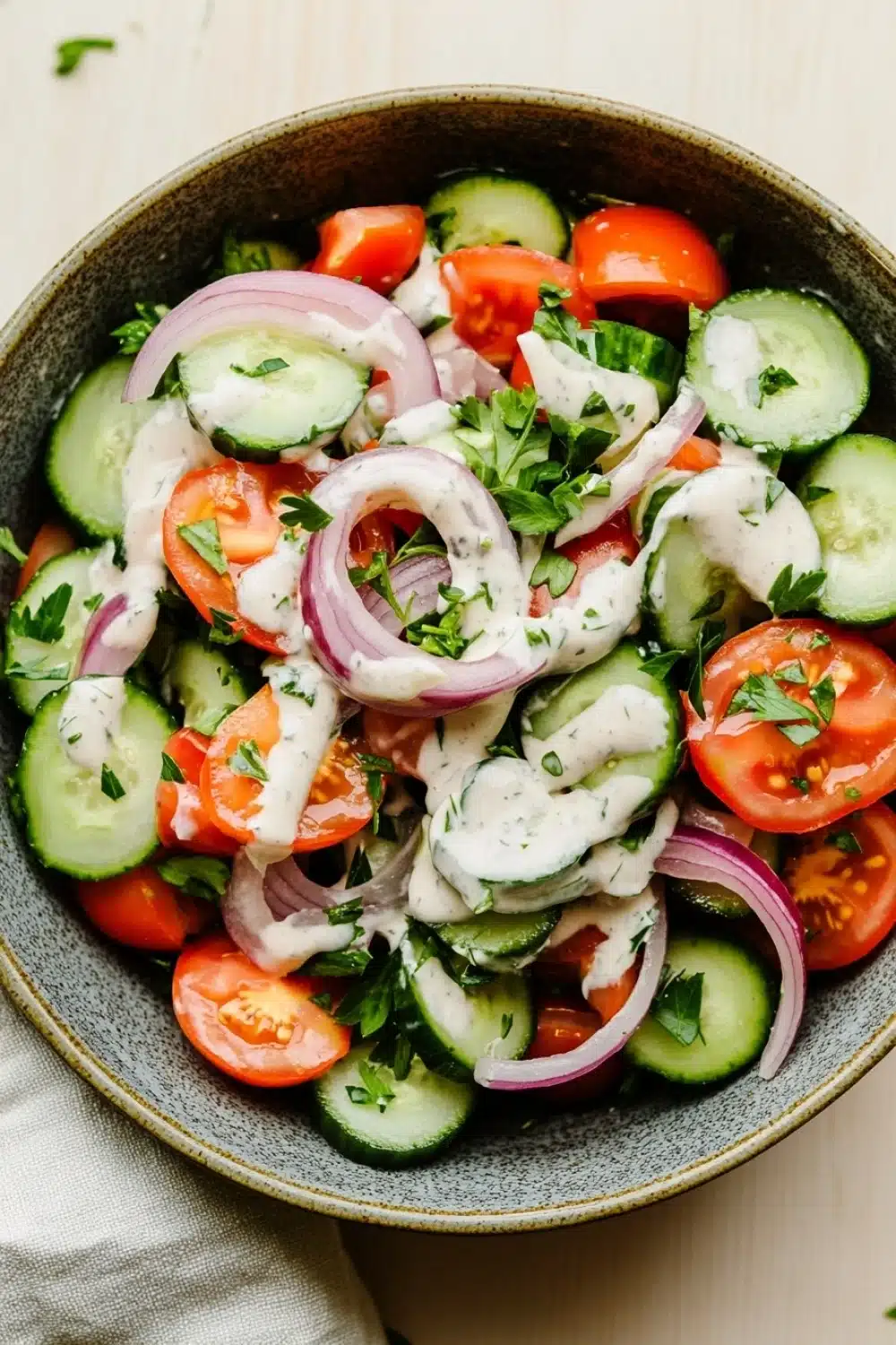 Cucumber tomato onion salad with ranch - the image is a close-up of a bowl of salad. the salad is made up of sliced cucumbers, red onions, cherry tomatoes, and cilantro. the cucumbers are cut into thin slices and are arranged in a circular pattern. the red onions are sliced into thin strips and are scattered throughout the salad. there is also a dollop of white dressing on top of the cucumbers. the bowl is sitting on a white tablecloth with a few sprigs of parsley scattered around it.