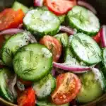Cucumber tomato onion salad with ranch - the image is a close-up of a bowl of salad. the salad is made up of sliced cucumbers, red onions, and cherry tomatoes. the cucumbers are cut into thin slices and are arranged in a circular pattern. the red onions are sliced into thin strips and are scattered throughout the salad. there are also some dill seeds sprinkled on top. the bowl is made of dark brown ceramic and is sitting on a wooden table. the background is blurred, but it appears to be a kitchen countertop.