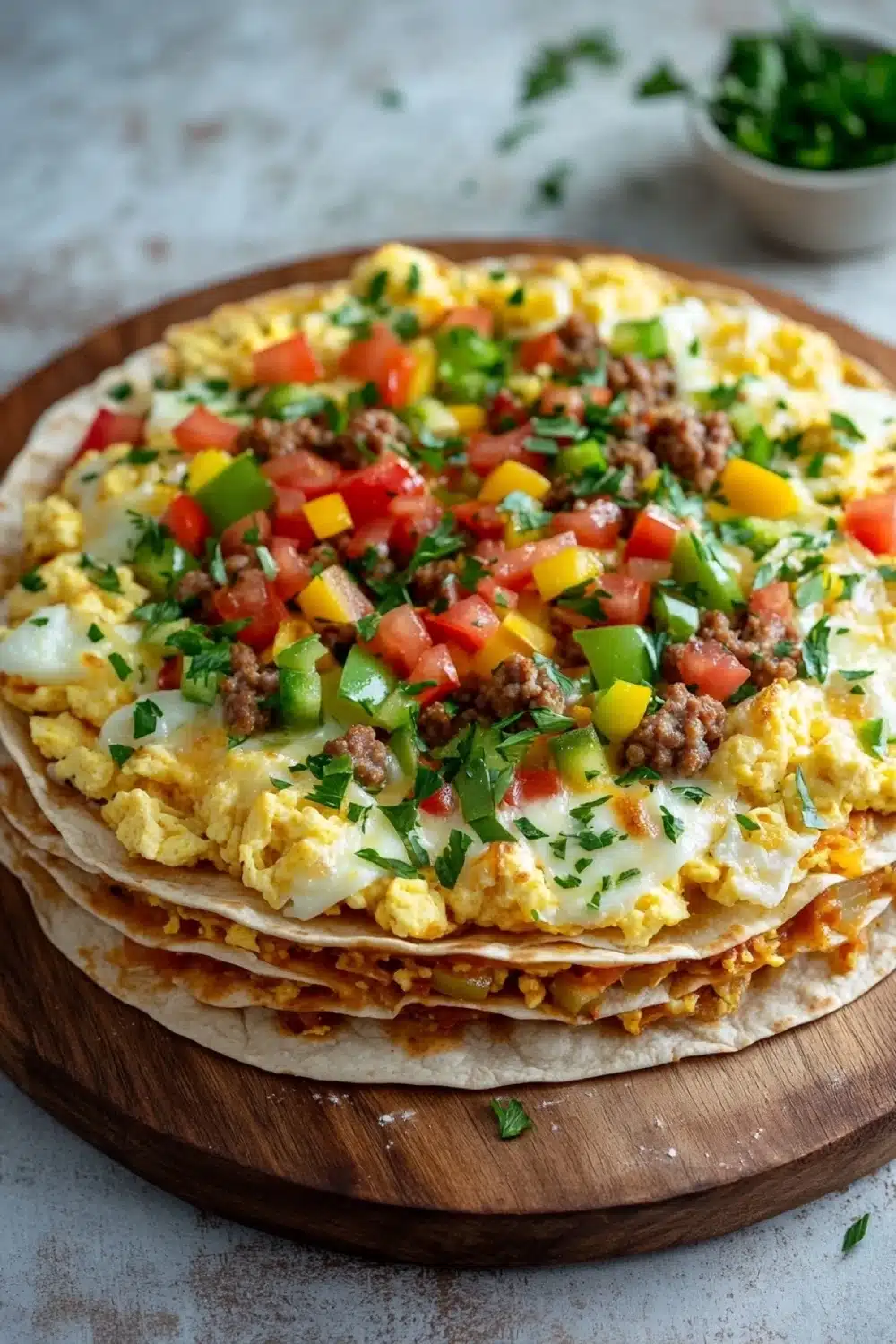 Deep dish breakfast pizza with tortillas - the image shows a stack of three flatbreads on a wooden cutting board. the flatbread is topped with scrambled eggs, diced tomatoes, diced bell peppers, and ground beef. the eggs are yellow and runny, and the toppings are arranged in a circular pattern. the toppings appear to be fresh and appetizing. in the background, there is a small bowl of chopped cilantro and a sprig of parsley. the cutting board is sitting on a white countertop.
