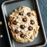 Edible cookie dough with oatmeal - the image shows a freshly baked cookie in a metal baking tray. the cookie appears to be oatmeal raisin cookies with chocolate chips scattered throughout. the tray is placed on a dark blue textured surface with scattered walnuts around it. there are also a few pieces of chocolate scattered around the tray, adding a pop of color to the image.