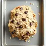 Edible cookie dough with oatmeal - the image shows a single cookie on a baking tray. the cookie appears to be freshly baked and has a golden brown color. it is topped with chocolate chips and oats, giving it a crunchy texture. the tray is lined with parchment paper and there are scattered oats around the cookie. the background is a white marble countertop.