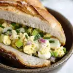 Egg salad sandwich baguette - the image shows a close-up of a sandwich in a bowl. the sandwich is made with a toasted baguette and is filled with scrambled eggs, red onions, and green onions. the eggs are yellow and runny, and the onions are a bright green color. the bowl is brown and has a textured surface. the background is blurred, but it appears to be a white marble countertop.