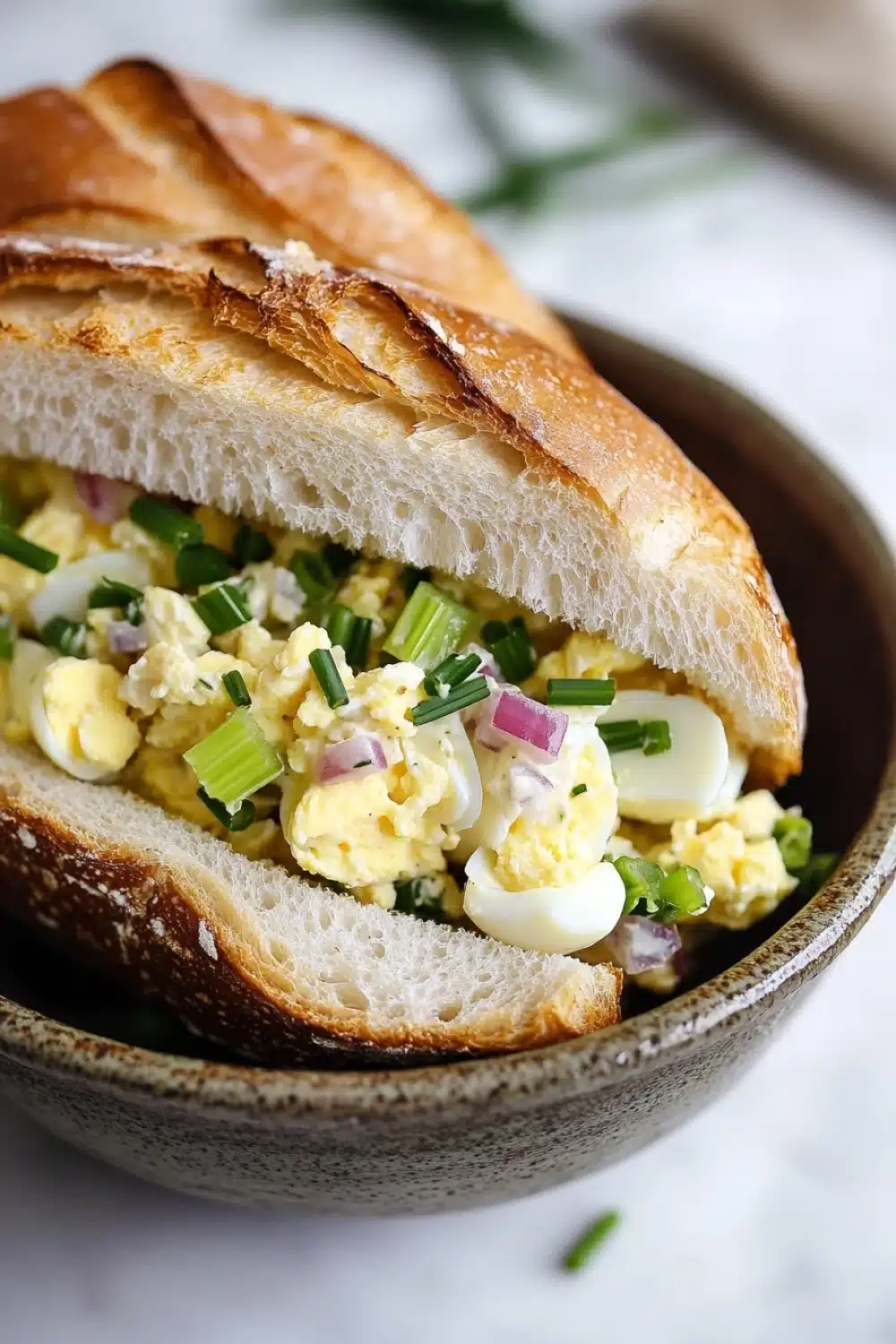 Egg salad sandwich baguette - the image shows a close-up of a sandwich in a bowl. the sandwich is made with a toasted baguette and is filled with scrambled eggs, red onions, and green onions. the eggs are yellow and runny, and the onions are a bright green color. the bowl is brown and has a textured surface. the background is blurred, but it appears to be a white marble countertop.