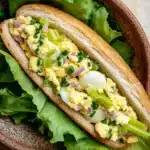 Egg salad sandwich baguette - the image shows a sandwich in a brown ceramic bowl. the sandwich is made with a long, toasted bun and is filled with scrambled eggs, diced onions, and green onions. the eggs are cooked sunny-side up and are arranged in a circular pattern on top of the scrambled eggs. the lettuce leaves are scattered around the sandwich, adding a pop of color to the dish. the bowl is sitting on a beige countertop.