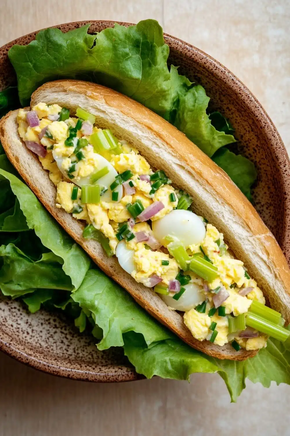 Egg salad sandwich baguette - the image shows a sandwich in a brown ceramic bowl. the sandwich is made with a long, toasted bun and is filled with scrambled eggs, diced onions, and green onions. the eggs are cooked sunny-side up and are arranged in a circular pattern on top of the scrambled eggs. the lettuce leaves are scattered around the sandwich, adding a pop of color to the dish. the bowl is sitting on a beige countertop.