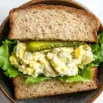Egg salad sandwich with dill pickles - the image shows a sandwich in a brown ceramic bowl. the sandwich is made with two slices of toasted bread and is filled with a mixture of scrambled eggs, lettuce, and pickles. the eggs are yellow and runny, and the pickles are green. the lettuce is a light green color and appears to be wilted. the bowl is sitting on a white marble countertop.