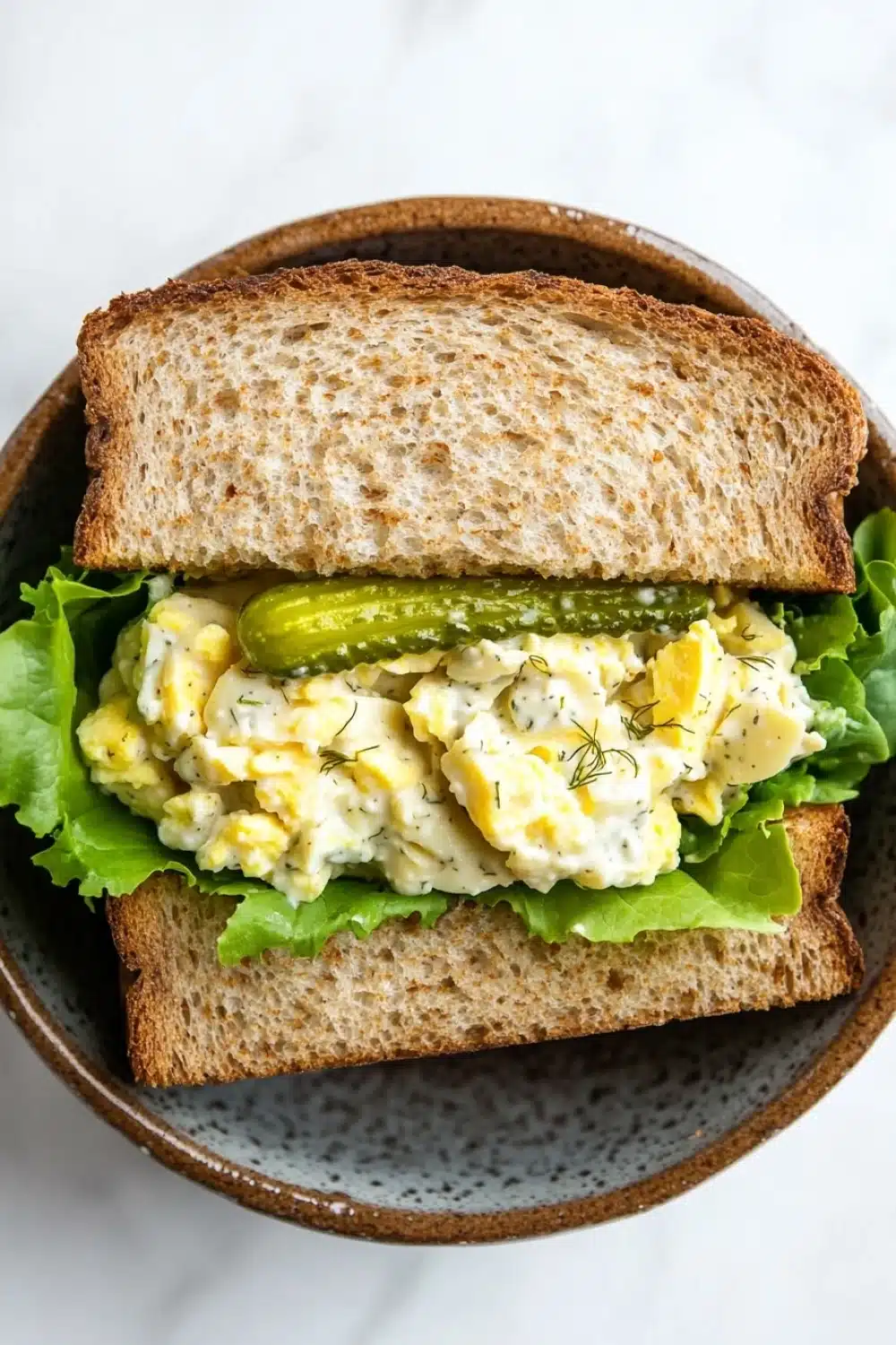 Egg salad sandwich with dill pickles - the image shows a sandwich in a brown ceramic bowl. the sandwich is made with two slices of toasted bread and is filled with a mixture of scrambled eggs, lettuce, and pickles. the eggs are yellow and runny, and the pickles are green. the lettuce is a light green color and appears to be wilted. the bowl is sitting on a white marble countertop.