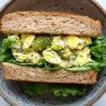 Egg salad sandwich with dill pickles - the image shows a sandwich in a brown ceramic bowl. the sandwich is made with two slices of toasted bread and is filled with a mixture of scrambled eggs, pickles, and green lettuce. the eggs are yellow and runny, and the pickles are green. the lettuce is a mix of green and yellow, and there is a sprinkle of dill on top. the bowl is sitting on a white marble countertop.