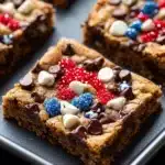 Fourth of july cookie bar - the image shows a tray of freshly baked brownies on a black countertop. the brownies are rectangular in shape and have a golden brown color. they are topped with chocolate chips, red, white, and blue sprinkles. the sprinkles are arranged in a circular pattern on top of the brownies. the tray is lined with parchment paper. the background is blurred, but it appears to be a kitchen countertop with more brownies scattered around.