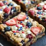Fourth of july cookie bar - the image shows a tray of freshly baked granola bars. the bars are golden brown and have a crumbly texture. they are topped with fresh strawberries, blueberries, and white chocolate chips. the berries are arranged in a circular pattern on top of the bars, creating a colorful and appetizing display. the tray is lined with parchment paper, and the background is dark, making the bars stand out.