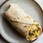 Freezer breakfast burritos with veggies - the image shows a tortilla wrap on a white marble plate. the wrap is rolled up and appears to be freshly made. it is filled with a mixture of scrambled eggs, vegetables, and herbs. the plate is round and has a light brown rim. the background is blurred, but it seems to be a kitchen countertop.