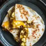 Freezer breakfast quesadilla - the image shows a stack of three quesadillas on a dark blue plate. the quesads are golden brown and appear to be freshly cooked. they are stacked on top of each other, with the top one slightly overlapping the bottom one. the top one is filled with scrambled eggs, sausage, and other ingredients. the plate is sitting on a wooden table.