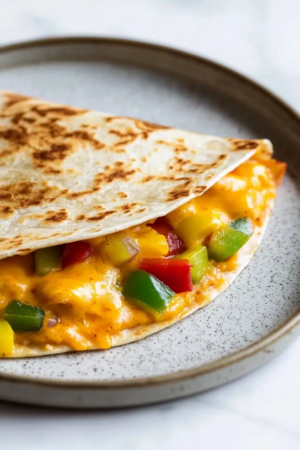 Freezer breakfast quesadilla - the image shows a quesadilla on a gray plate with a white speckled rim. the tortilla is golden brown and appears to be freshly cooked. it is filled with a mixture of yellow cheese, red and green bell peppers, and other vegetables. the cheese is melted and bubbly, and the vegetables are arranged in a circular pattern around the edges of the tortilla. the plate is sitting on a white marble countertop.