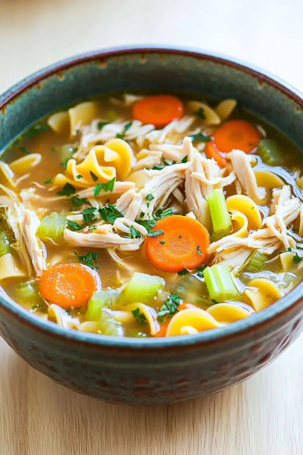 Freezer chicken noodle soup - the image is a close-up of a bowl of soup. the soup appears to be a type of noodle soup with chunks of chicken, carrots, celery, and other vegetables mixed in. the bowl is made of ceramic and has a dark blue rim. the broth is a light brown color and looks thick and creamy. there are also some chopped herbs and spices scattered throughout the soup, adding a pop of green color to the dish. the background is a wooden table, and the bowl is sitting on a white surface.
