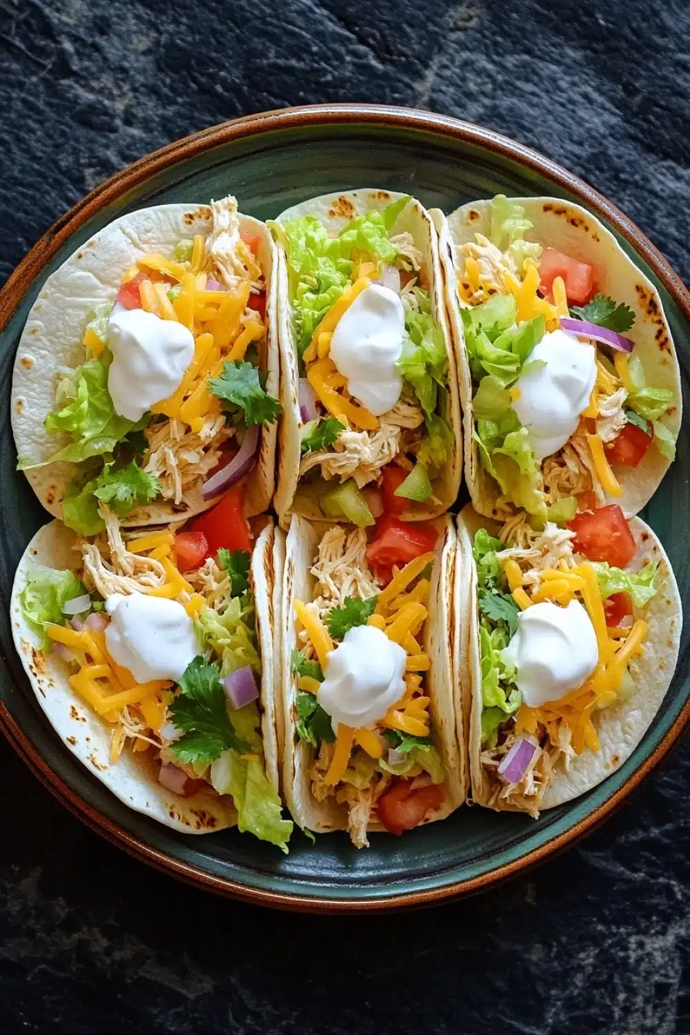 Freezer chicken tacos - the image shows four tacos arranged in a circular pattern on a green plate. the tacos are filled with shredded chicken, shredded cheese, diced tomatoes, red onions, and cilantro. each taco is topped with a dollop of sour cream. the plate is sitting on a dark textured surface.