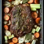 French roast beef slow cooker - the image shows a rectangular baking dish with a large piece of cooked meat on top. the meat appears to be a roast beef, with a dark brown color and a crispy texture. it is garnished with a sprig of fresh herbs, possibly thyme or rosemary. the dish is served on a bed of sliced carrots, celery, and onions. the vegetables are arranged in a way that they are overlapping each other, creating a colorful and appetizing presentation. the baking dish is lined with parchment paper, and the background is a dark grey countertop.