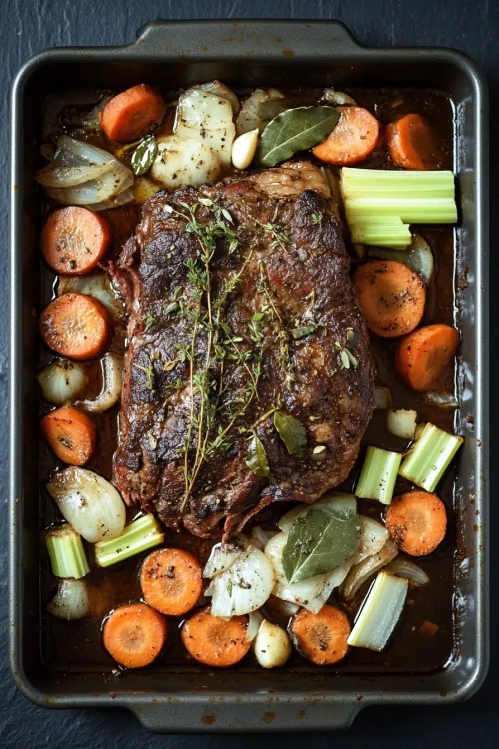 French roast beef slow cooker - the image shows a rectangular baking dish with a large piece of cooked meat on top. the meat appears to be a roast beef, with a dark brown color and a crispy texture. it is garnished with a sprig of fresh herbs, possibly thyme or rosemary. the dish is served on a bed of sliced carrots, celery, and onions. the vegetables are arranged in a way that they are overlapping each other, creating a colorful and appetizing presentation. the baking dish is lined with parchment paper, and the background is a dark grey countertop.
